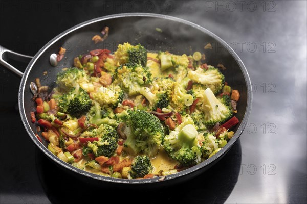 Vegetarian curry with broccoli and other vegetables in a steaming frying pan on a black stovetop,