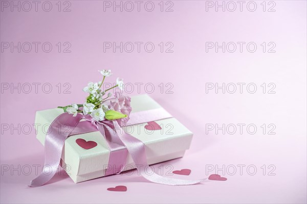 Gift box with flower bouquet, ribbon and paper heart shapes on a light pink background, love