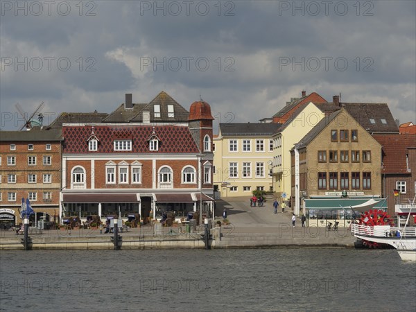 Historic buildings and tourists along a harbour shore under a cloudy sky, Kappeln,