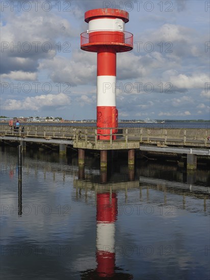 Lighthouse in red and white at the pier, reflecting on calm water under partly cloudy sky,