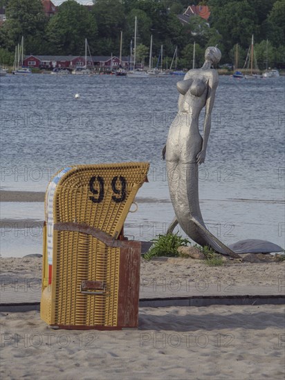 A single beach chair and a statue on a quiet sandy beach overlooking the water under a clear sky,