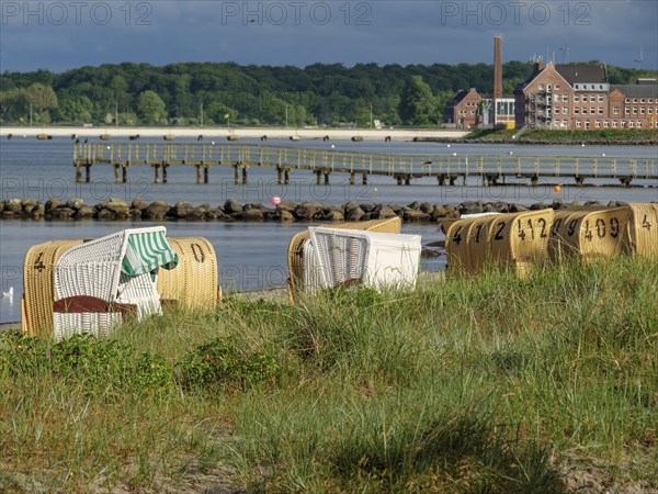 Some beach chairs stand on a grassy strip of dunes on the sandy beach, with a view of the water and