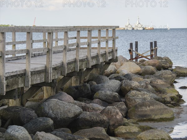 Wooden jetty next to large rocks on the shore with ships in the background, Eckernförde,