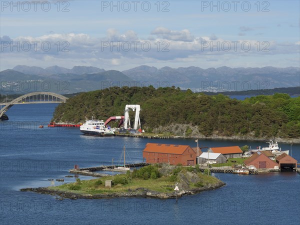 Small island with red buildings and boats, surrounded by the sea. A bridge and mountains are
