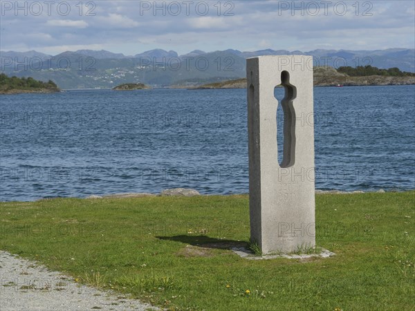 Concrete sculpture with a view of water and mountains, surrounded by green grass and a path in the