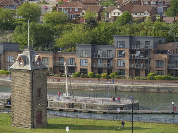 Stone tower on the riverside with modern residential buildings in the background, people walking on