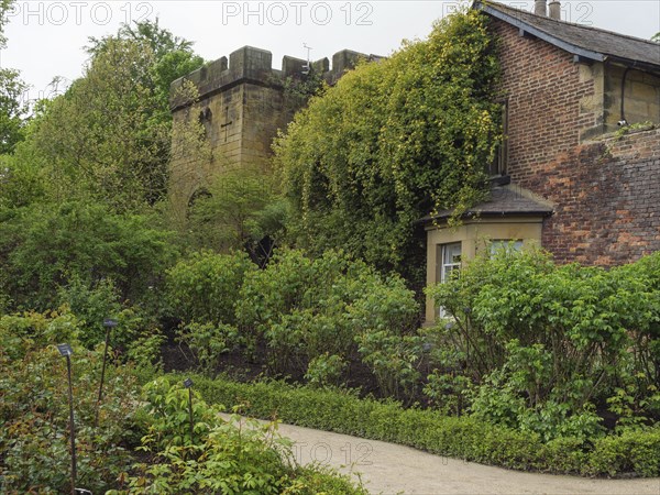 An old fortified tower and neighbouring building surrounded by dense greenery and flowers in a