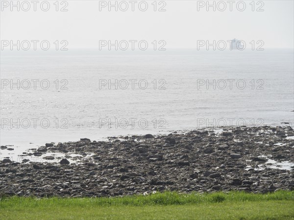 View of a rocky coastline merging into the open sea, a ship can be seen on the horizon in the fog,