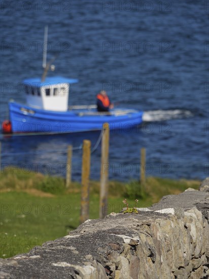 A blurred fishing boat and fence are seen behind a stone wall over blue water, Lerwick, Shetlands,