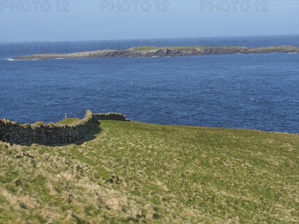 Green meadow with stone wall on the shore that merges into the wide blue sea and visible islands,