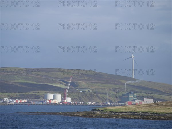 Coastal landscape with green hills, wind turbine and industrial buildings on the horizon, Lerwick,