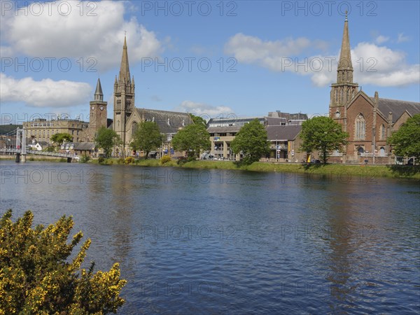 A cosy riverside town with striking church buildings and trees under a partly cloudy sky,