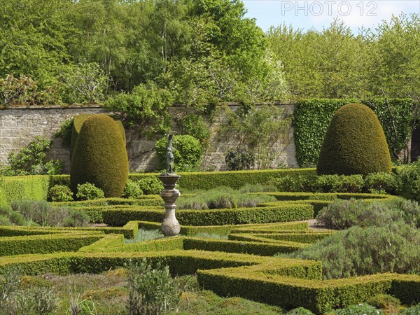 A manicured garden with symmetrical hedges and a centrally placed sculpture in a green landscape,