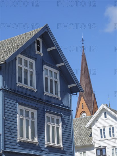 Blue wooden house with white windows in front of a red church tower under a bright blue sky,