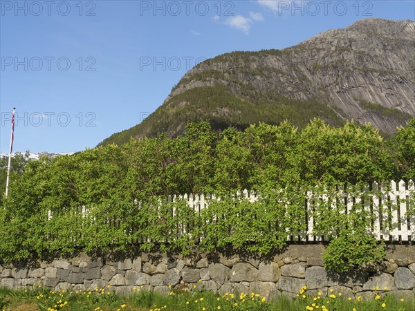 A stone wall with a white fence and green surroundings in front of a mountain under a clear sky,