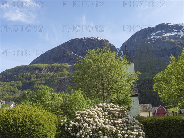A church behind blossoming trees and bushes with towering mountains in the background, Eidfjord NOR
