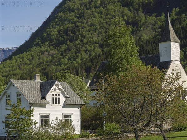 White houses and church in a village surrounded by green trees and mountains under a clear sky,