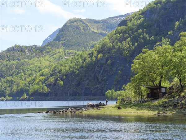 A small cabin on the shore of a clear lake, surrounded by mountains and dense forest, quiet and