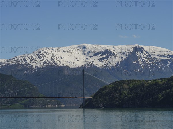 Suspension bridge over a fjord with snow-capped mountains in the background, blue sky and calm