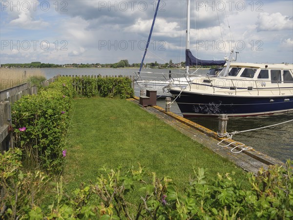 A sailing boat moored to a wooden jetty, surrounded by bushes and a cloudy sky, Arnis,