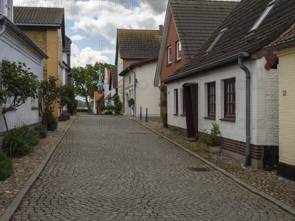 A quiet cobbled street with stylish houses and trees under a cloudy sky, Maasholm,
