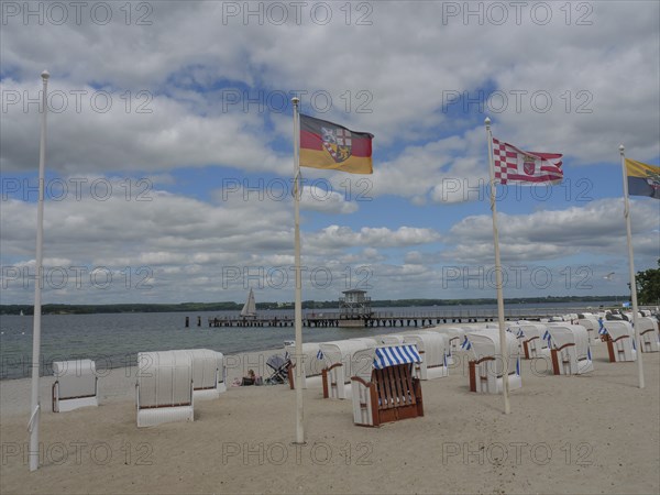 Beach chairs on a sandy beach under a cloudy sky, flags blowing in the wind, sea and jetty in the