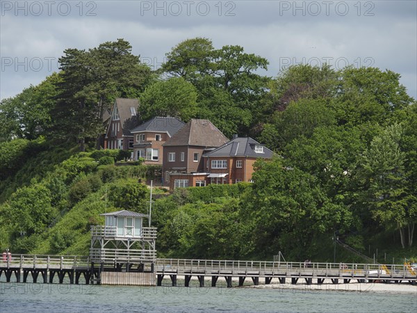 Houses on a green hill, surrounded by trees, with a footbridge leading into the water, under a