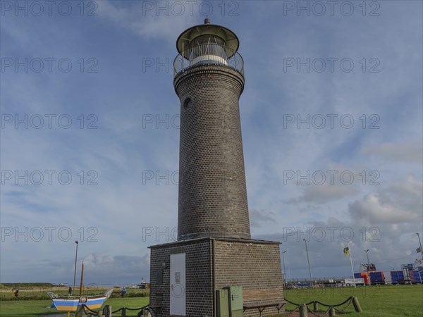 A tall brick lighthouse in a meadow with a few boats and a cloudy sky in the background, juist,