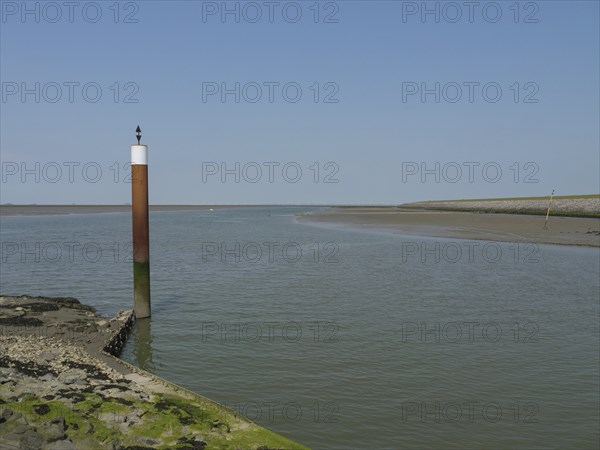A coastal scene with a single post in calm water with clear sky and rocky shore, Hallig Hooge,