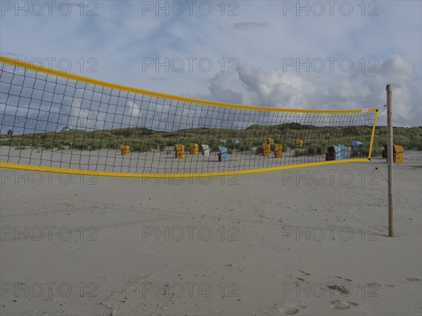 A yellow volleyball net on the beach with several beach chairs and dunes under a cloudy sky, juist,