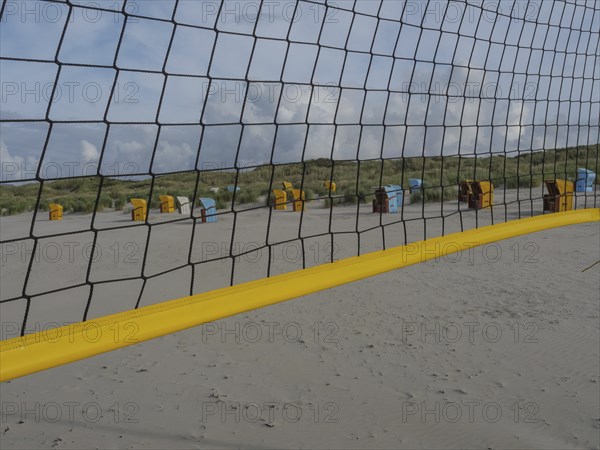 A volleyball net stretches across the beach, with dunes and beach chairs in the background, juist,