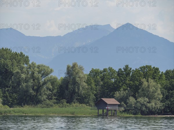 Small wooden hut on the shore of a lake, surrounded by trees with a mountain backdrop, Lindau, Lake