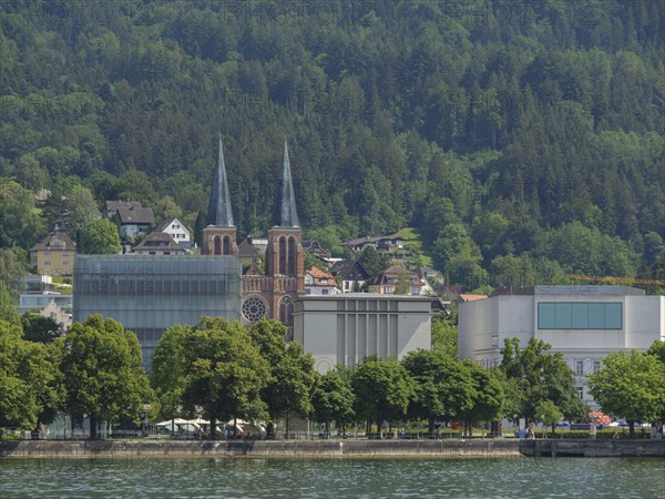 Two striking church towers and modern buildings in a green, wooded landscape on the lakeshore,