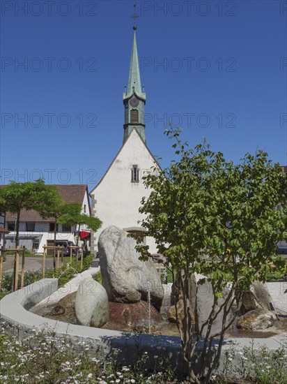 Church tower and fountain in a village surrounded by green trees and flowering plants on a sunny