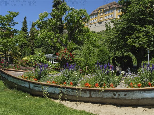 An old boat serving as a flower box in a green garden with a large yellow building in the