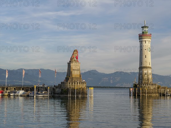 View of a harbour with a lion sculpture and a lighthouse against a mountain backdrop in the morning