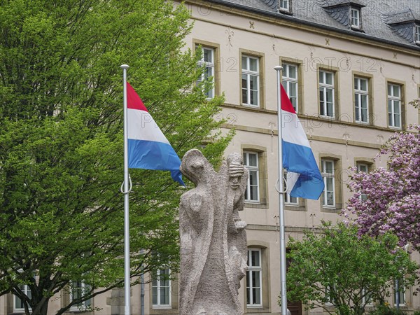 Monument with two Luxembourg flags in front of a historic building with blossoming trees,