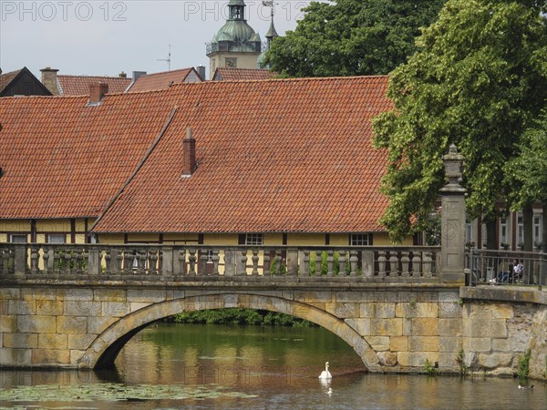 Stone bridge over a pond with red tiled buildings in the background and a duck in the water,