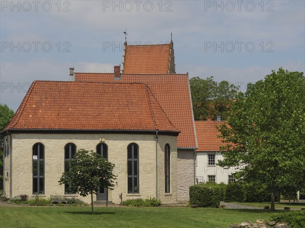A small chapel with red roof tiles and large windows, surrounded by meadows and a tree, under a