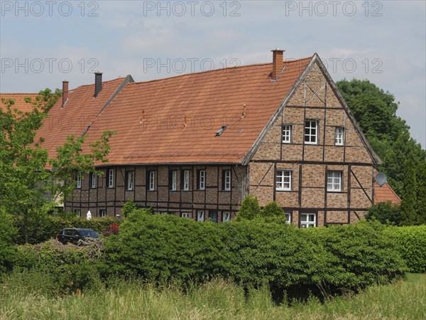 Large half-timbered house with red tiled roof surrounded by green bushes and trees on a summer's