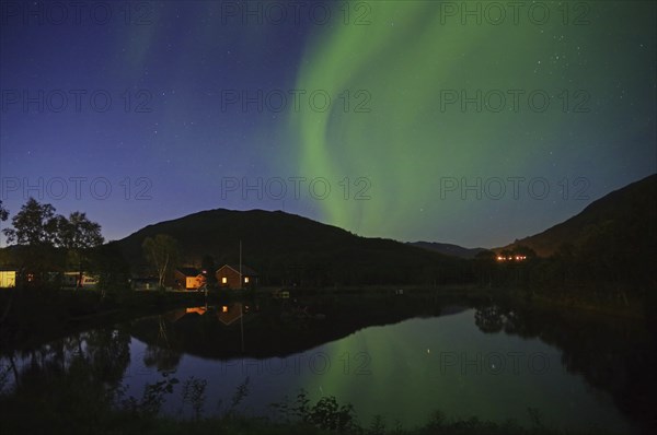 House and hill under the night sky with green northern lights and a reflection in the lake,