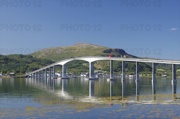 A curved bridge spans calm waters with reflective mountains and green landscapes under a clear sky,