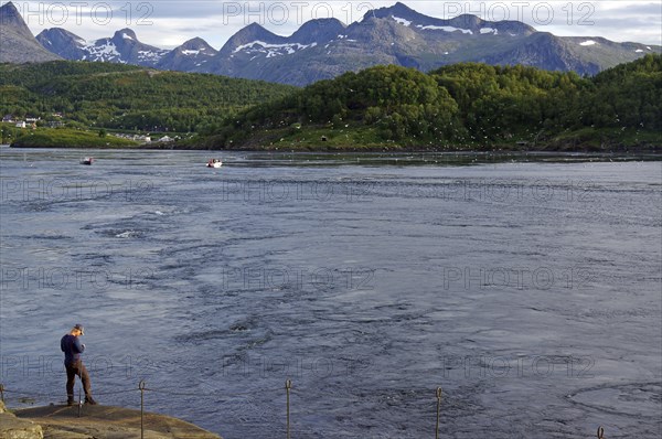 A man stands on the bank of a tidal stream, surrounded by green hills and mountains in the