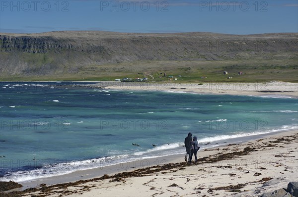 Two people walking along a beach with clear blue water and rocks in the background, Latrabjarg,