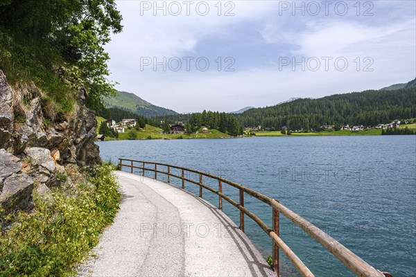 Path with railing along a lake, surrounded by mountains and forests under a slightly cloudy sky,