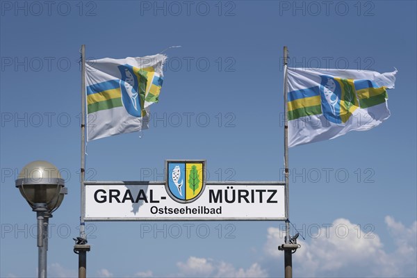 Two flags and a sign with the inscription Graal-Müritz Ostseeheilbad under a clear blue sky with