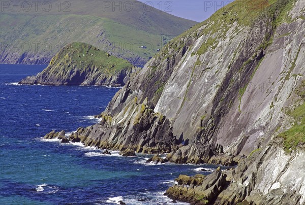 Breathtaking cliff landscape, coloured in green and grey, lined with deep blue sea and green hills,