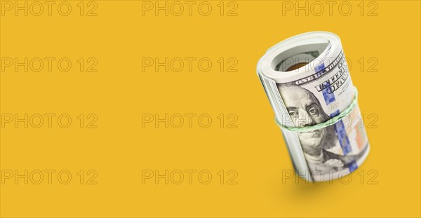 Roll of one hundred dollar bills secured with a rubber band on a yellow ochre background