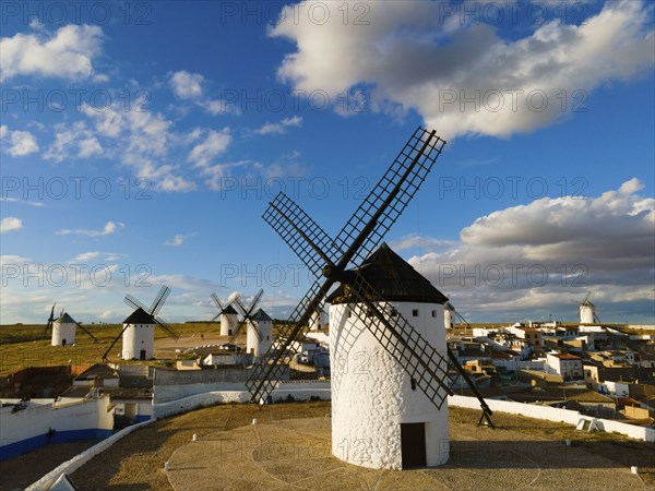 White windmills stand under a partly cloudy blue sky, with a village in the background, aerial