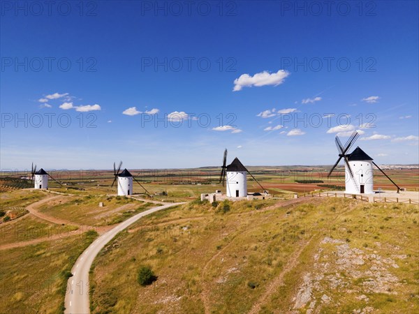 Several white windmills stand along a dirt road under a clear blue sky, aerial view, Alcazar de San